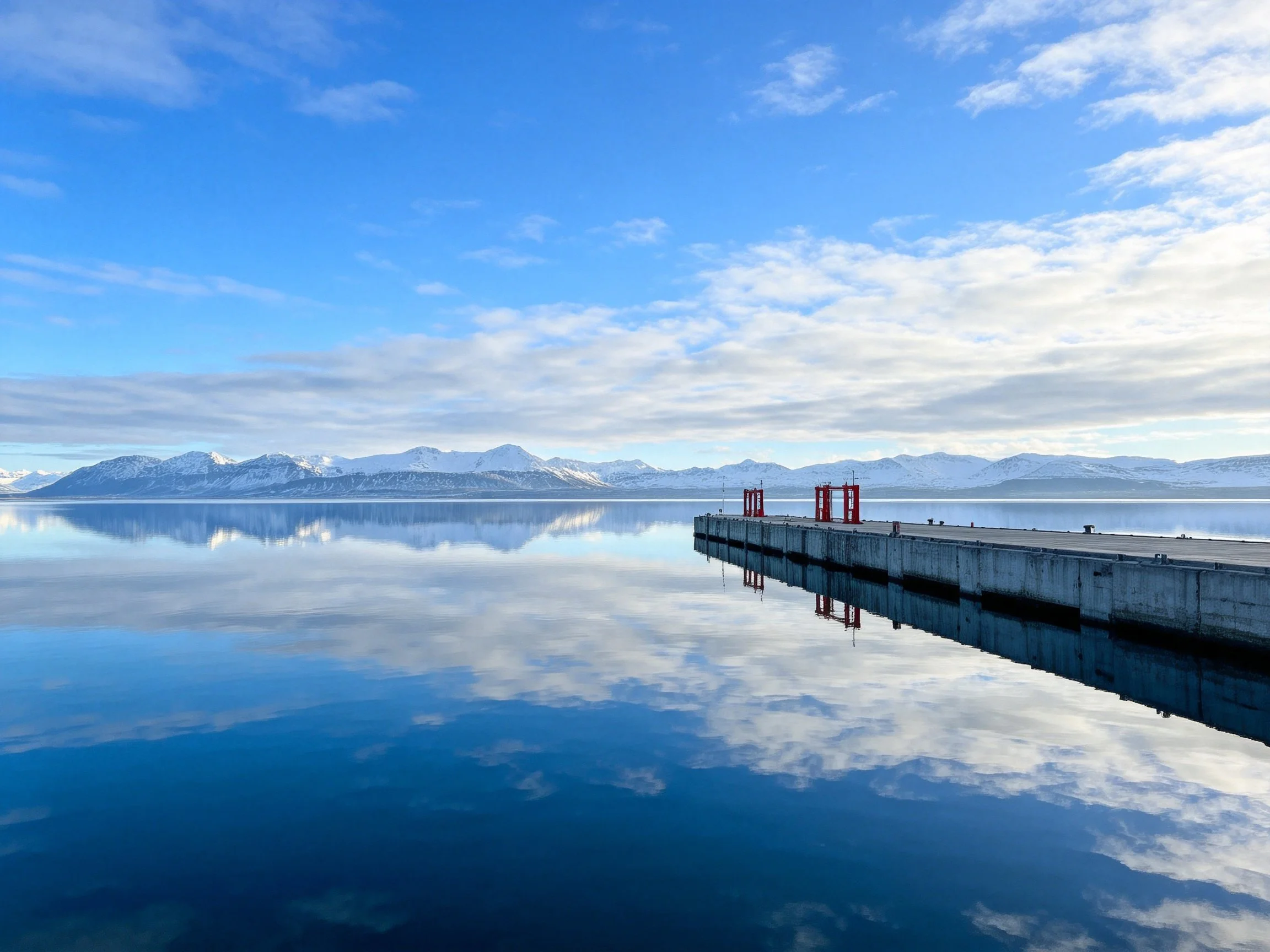 Concrete dock extends over calm lake reflecting snowy mountains