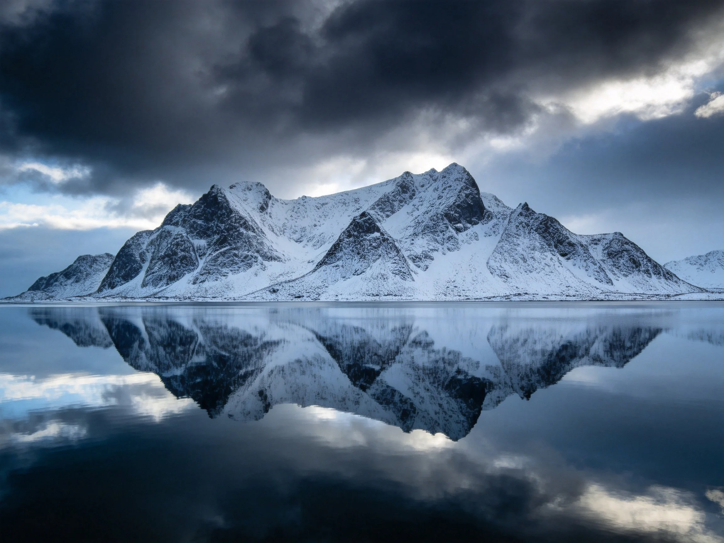 Dramatic snowy mountain range reflected in mirror-like waters under stormy skies
