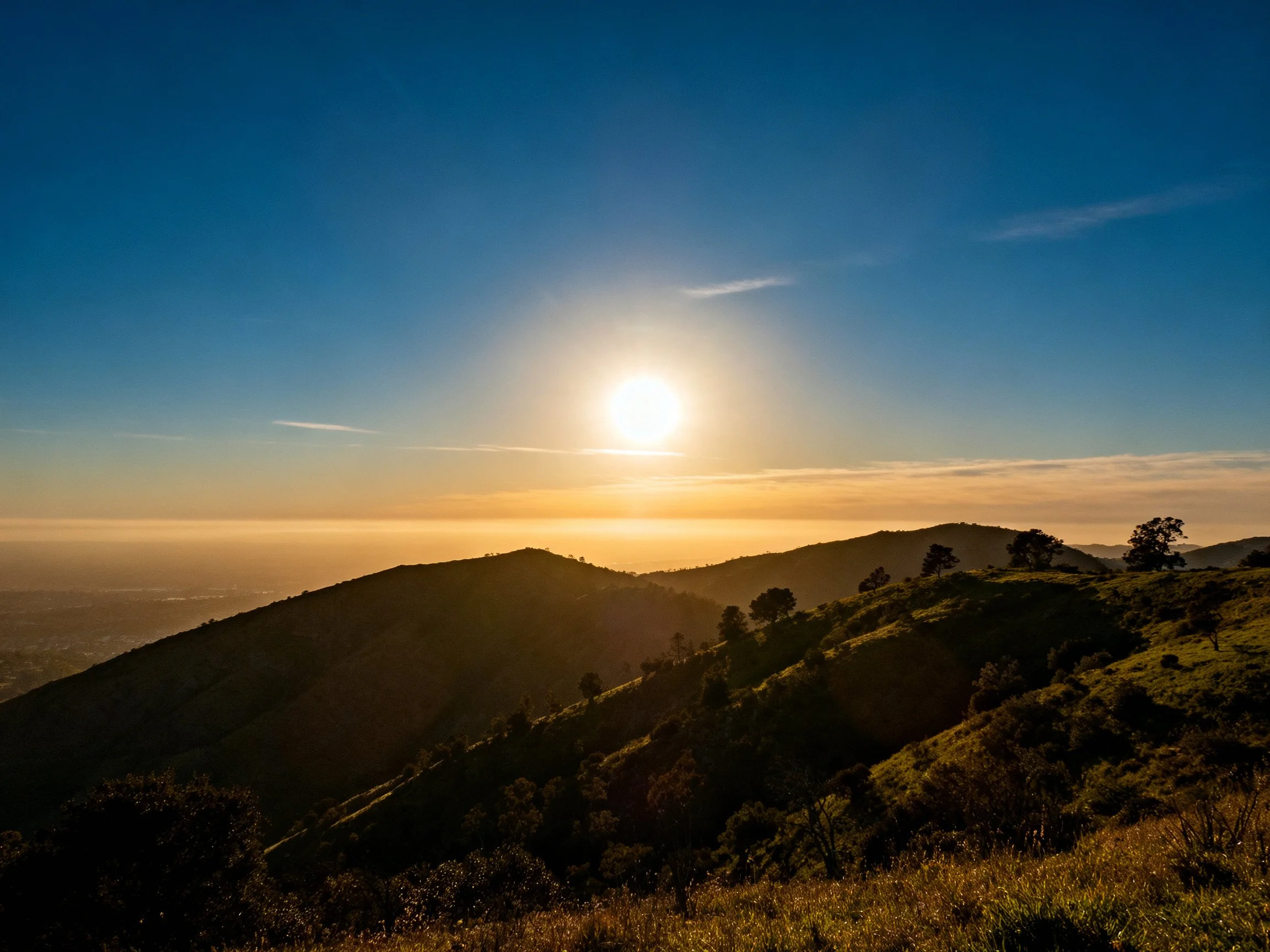 Golden sunrise illuminates rolling green hills under vast blue sky.