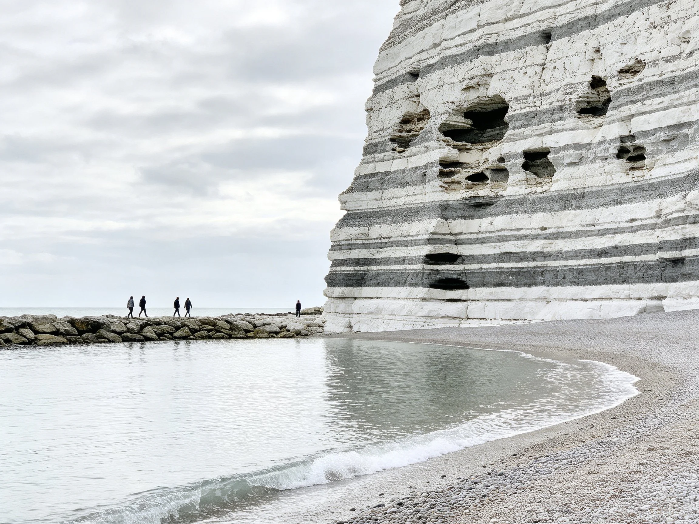 Layered white and gray cliffs tower over a pebbly beach with calm waters and distant walkers