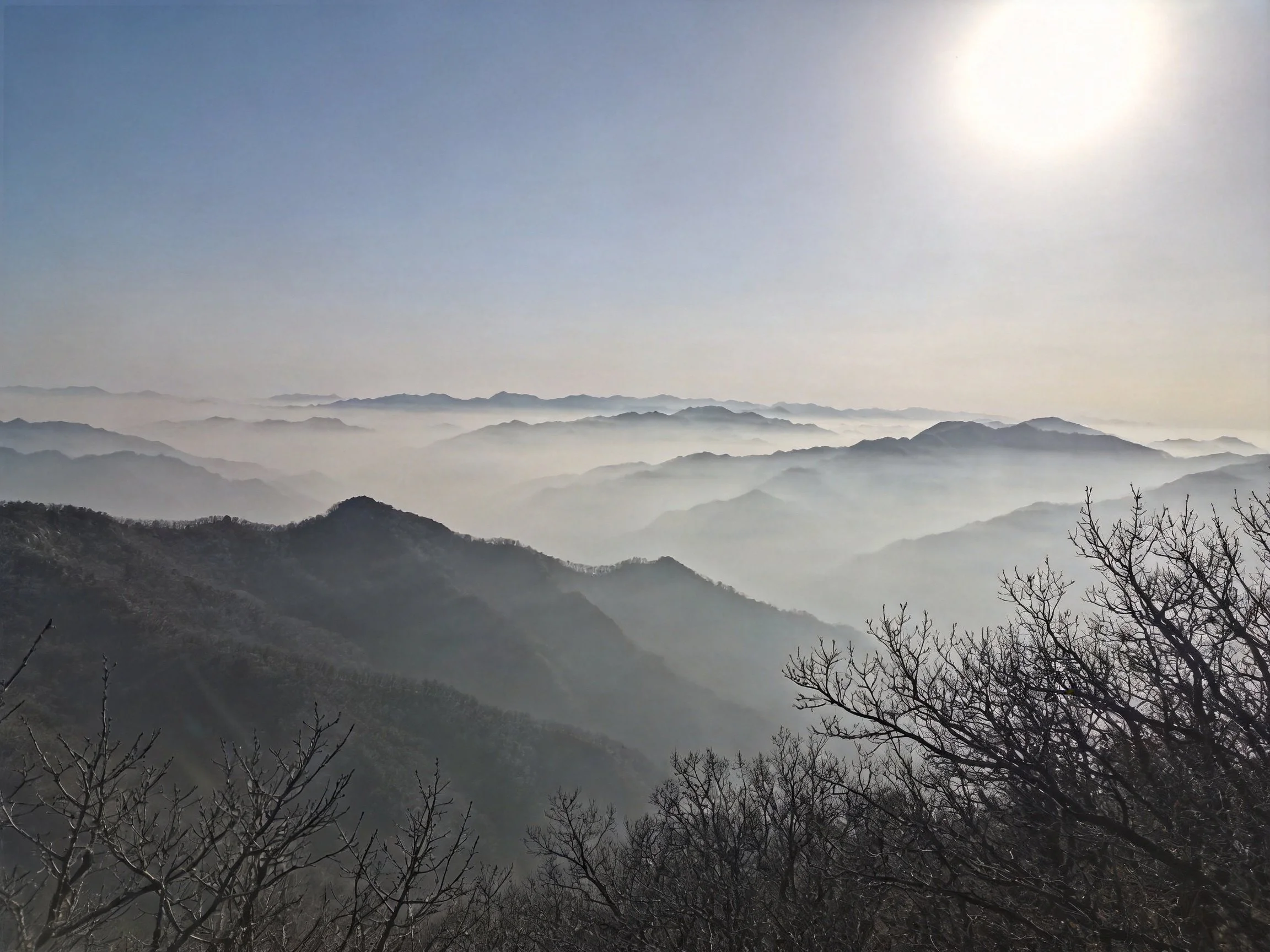 Misty mountain ridges extend into multiple foggy valleys with a bright sun overhead.