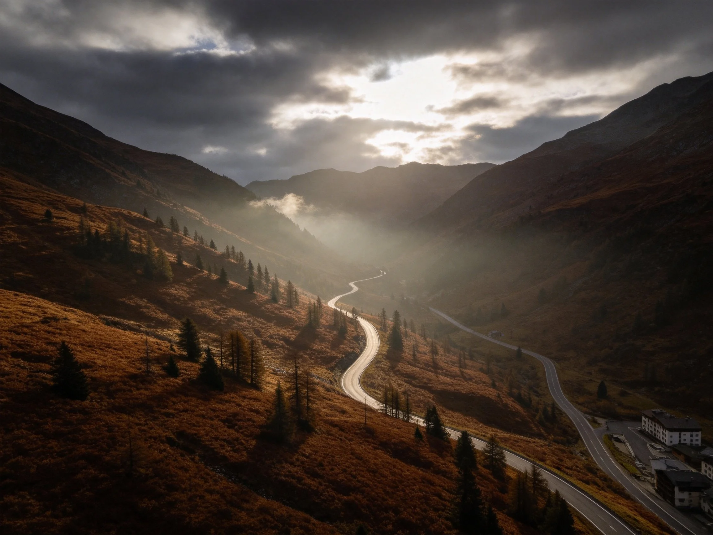 Mountain valley with winding roads, golden foliage, and dramatic cloudy sky.
