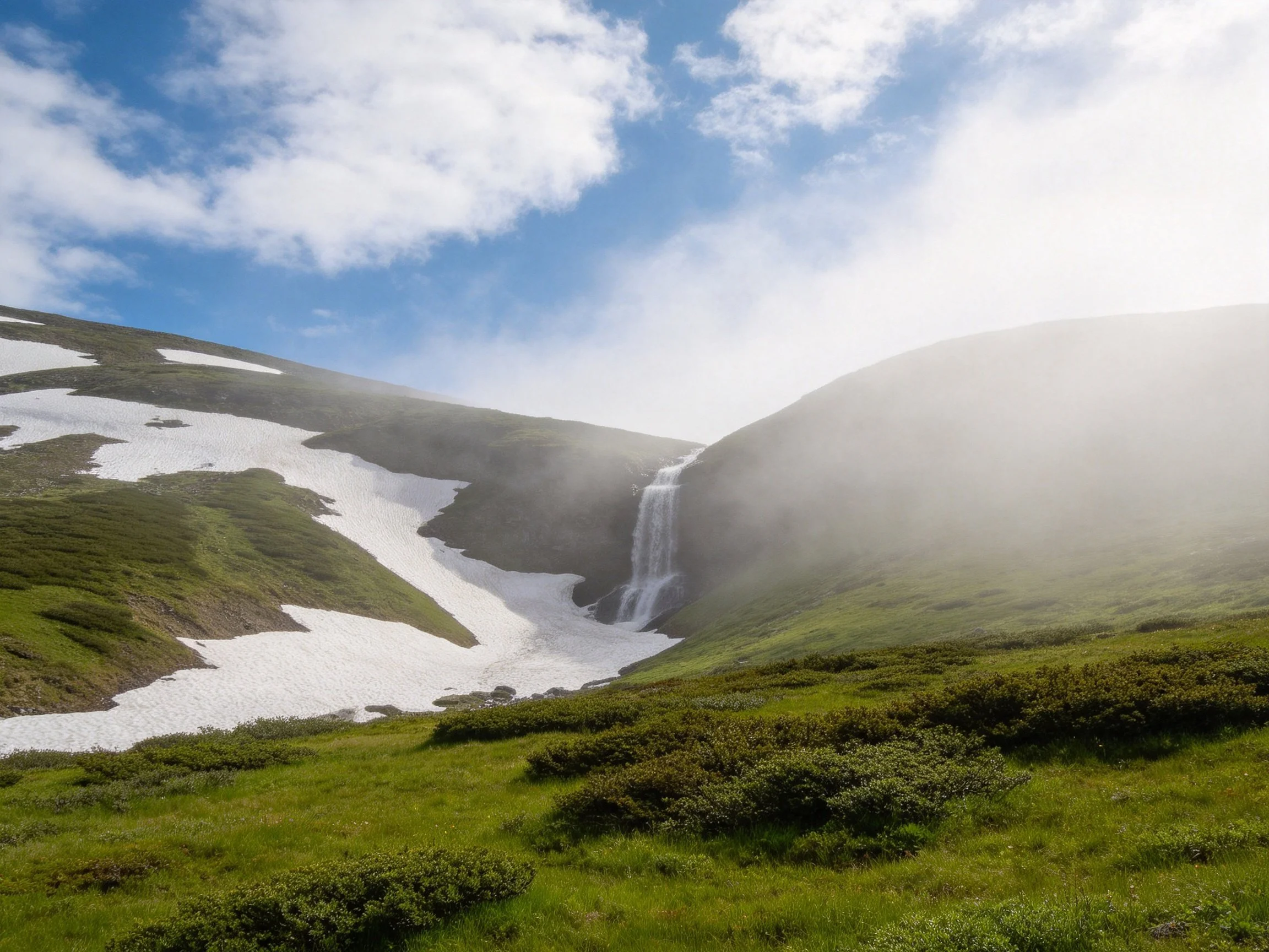 Mountain waterfall with snow patches and lush green terrain
