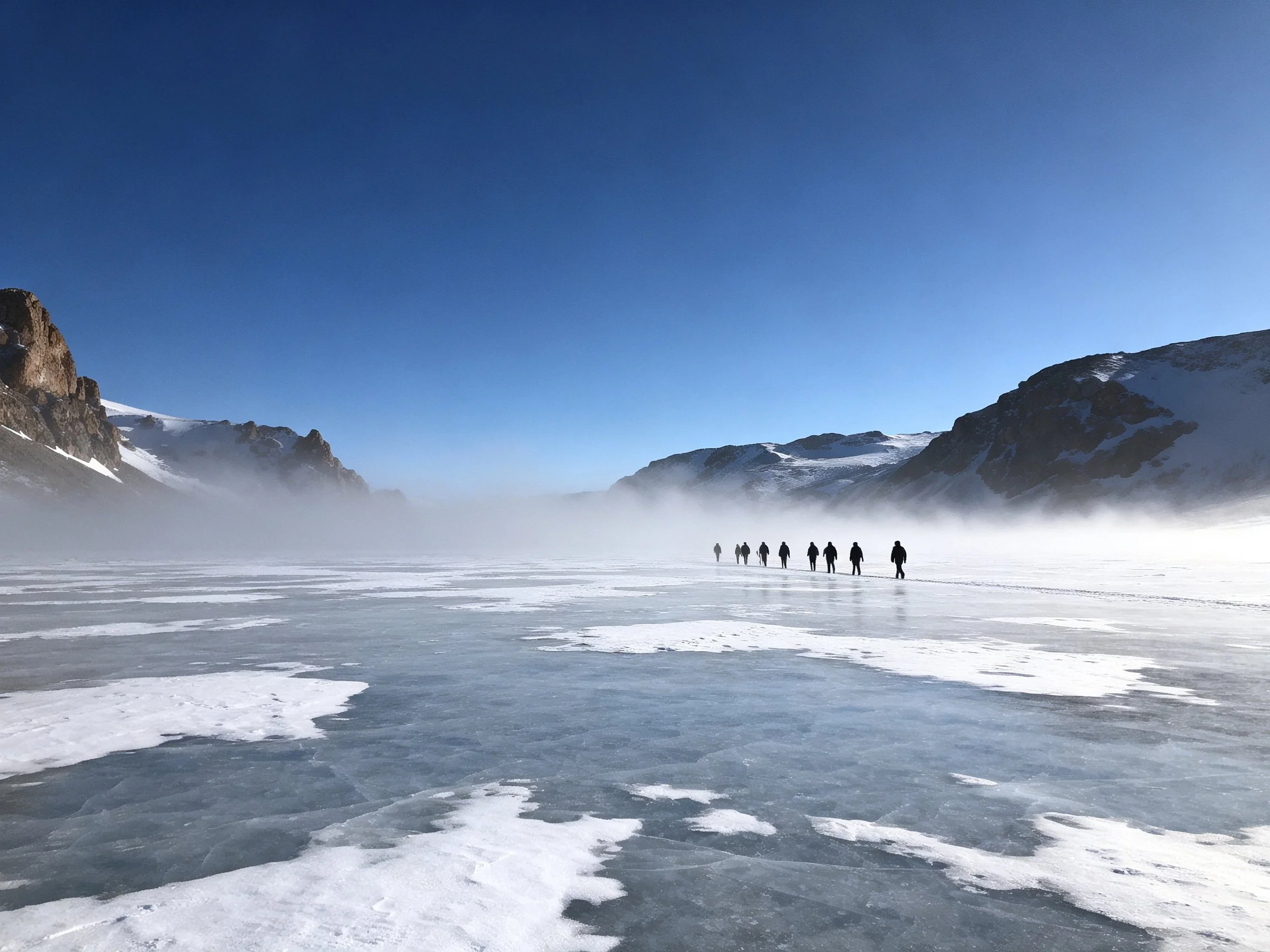 People walk across frozen terrain near snow - capped mountains under clear blue sky