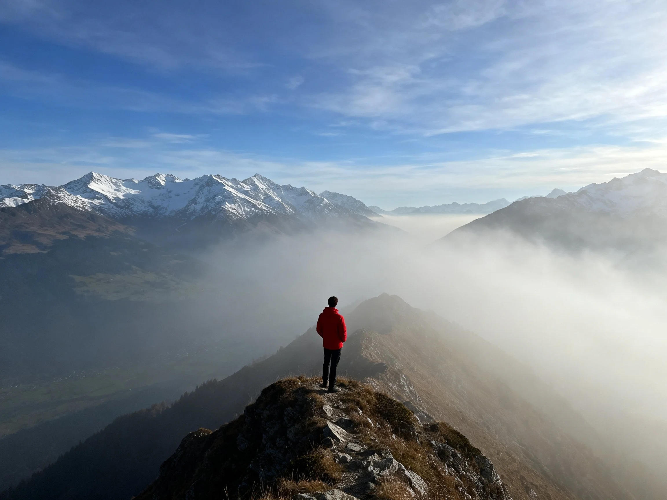 Person in bright red jacket atop rocky mountain ridge with snow-capped peaks and foggy valley