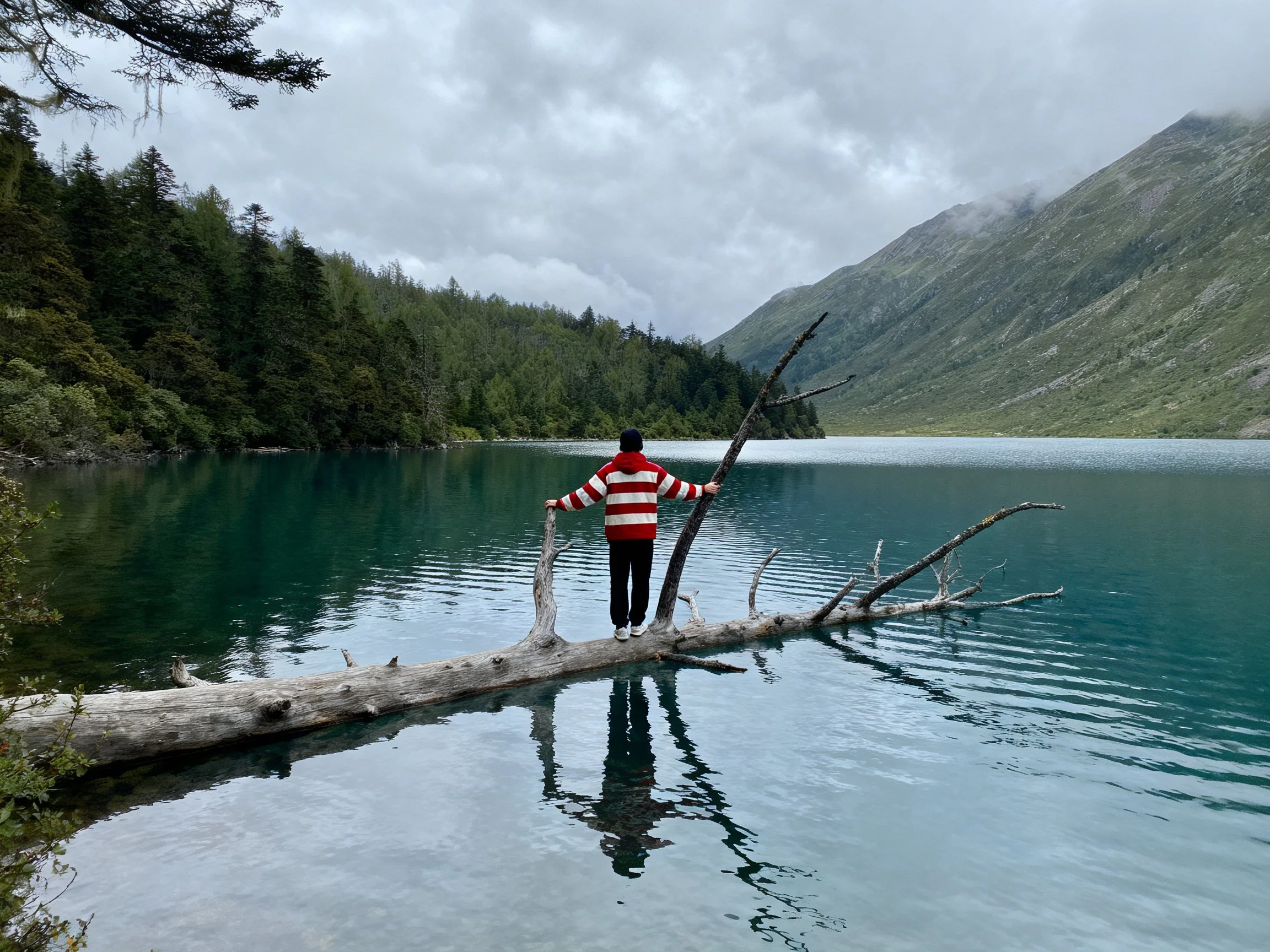 Person in striped hoodie on log over turquoise lake near forested mountains