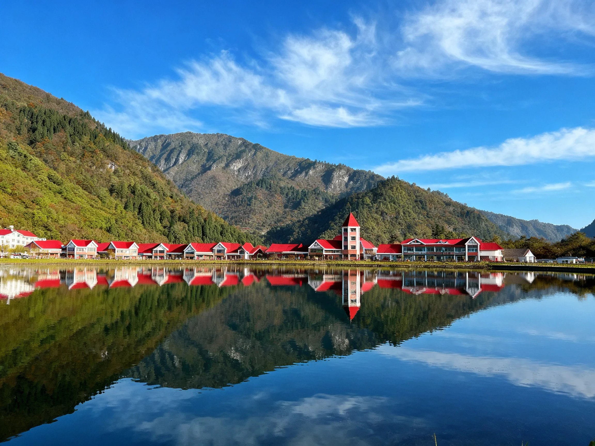 Red-roofed buildings reflected in calm lake amidst mountainous terrain