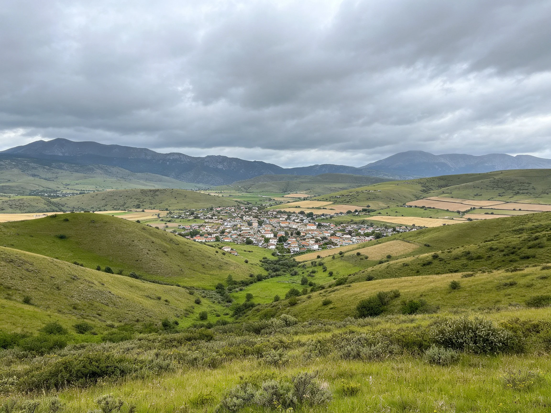 Rolling green hills encircle a village under stormy mountain skies.