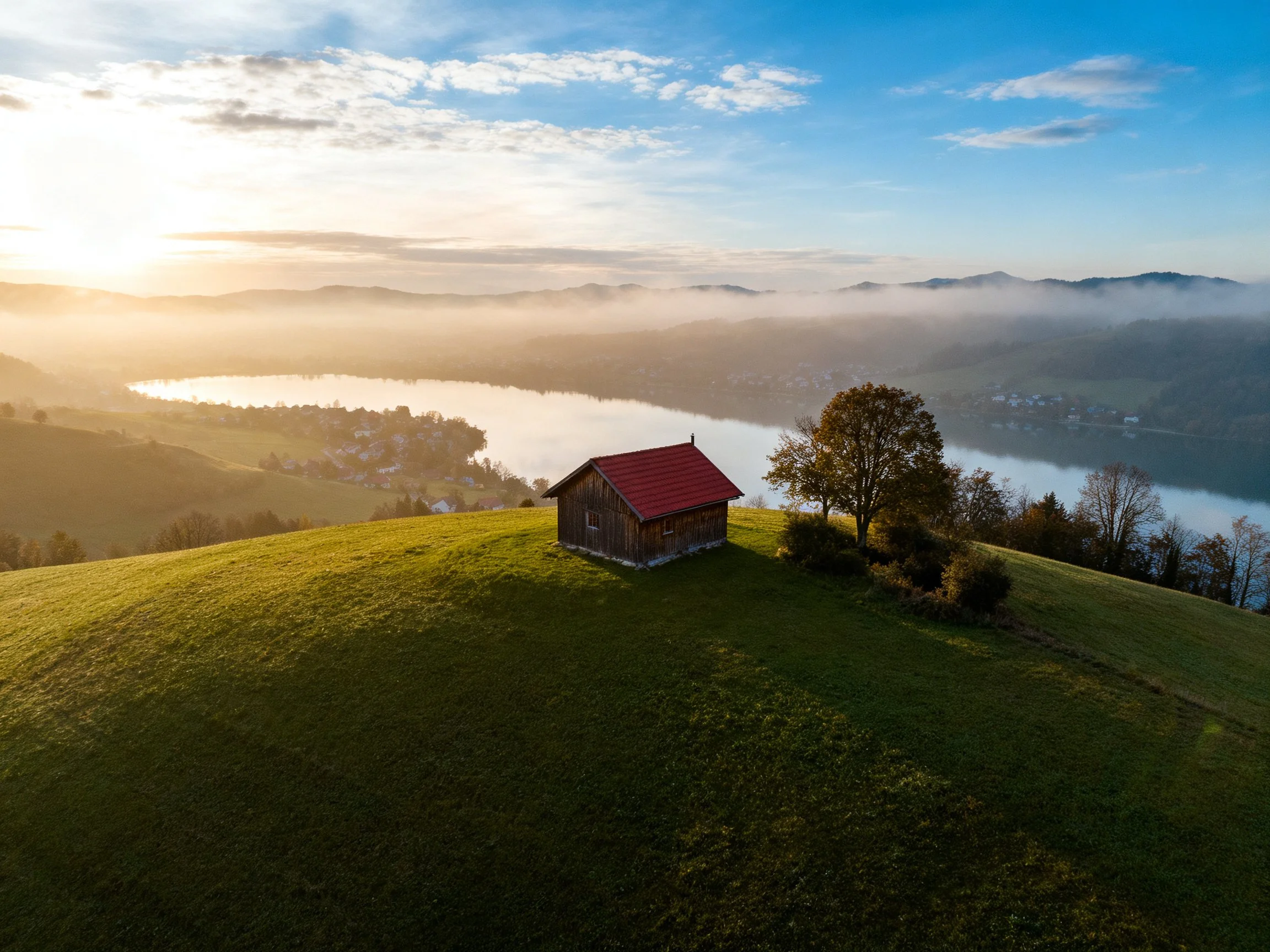 Rustic cabin with red roof on green hill near misty lake