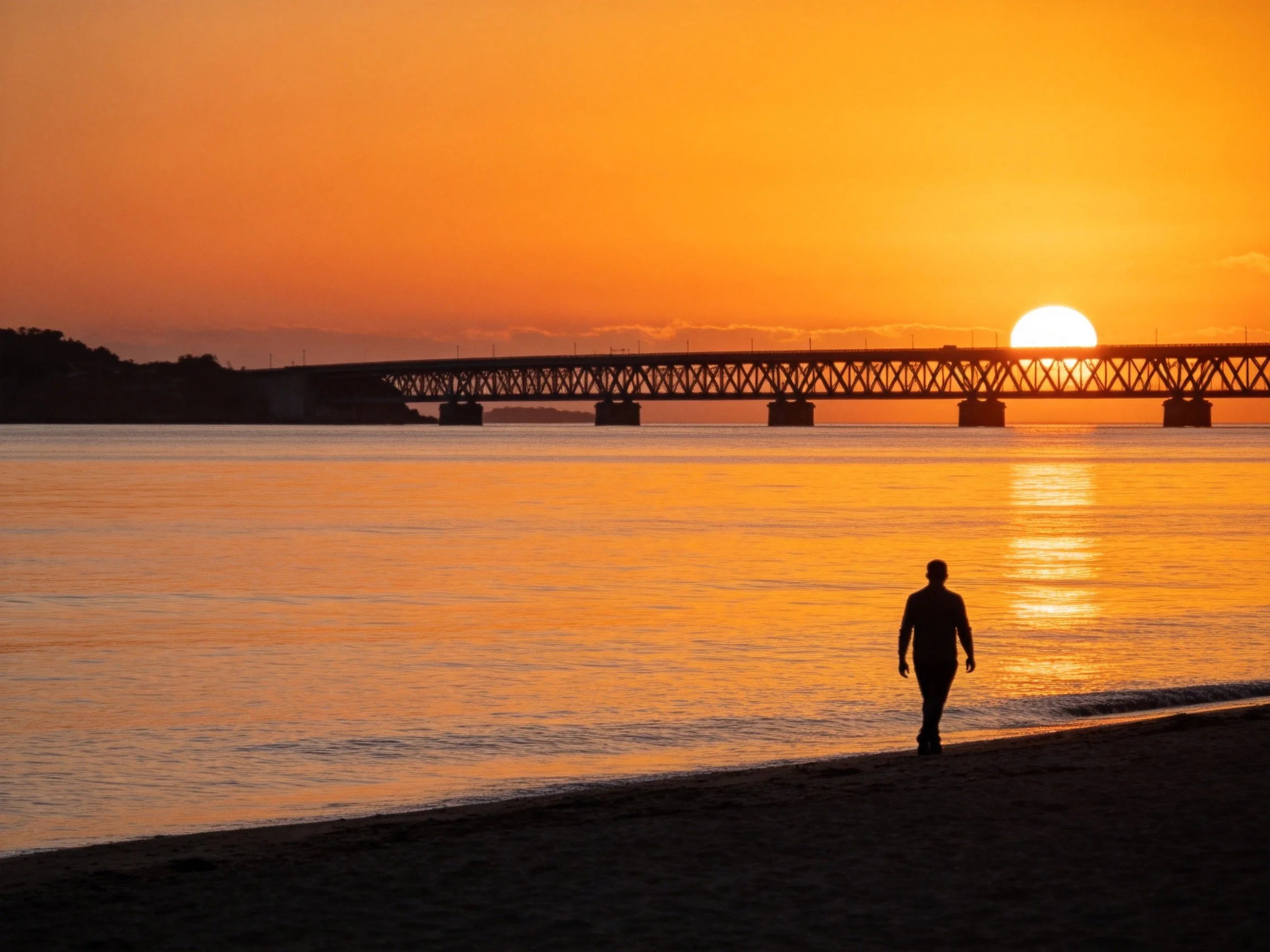 Silhouette of person walking along beach during vivid orange sunset with bridge