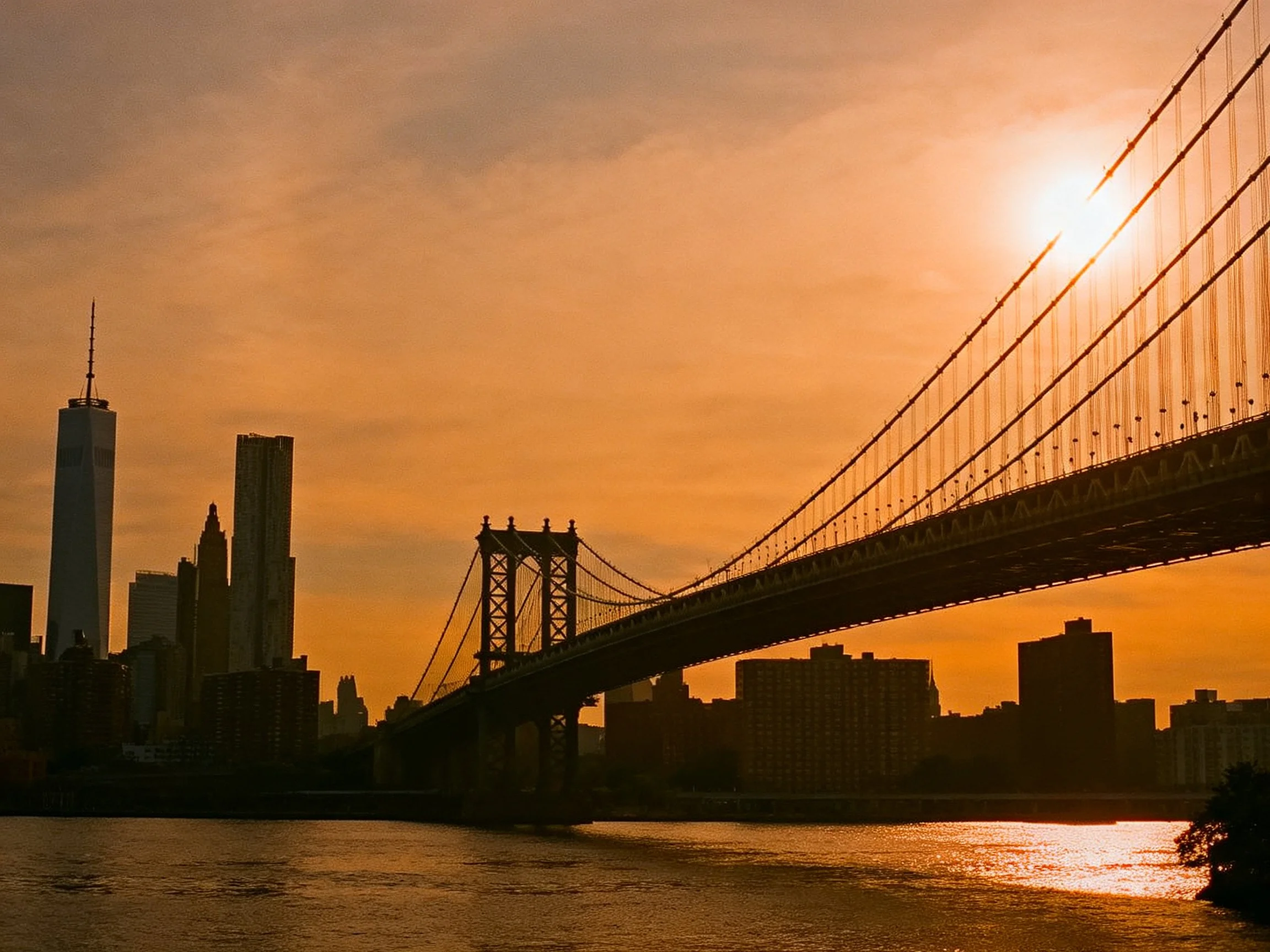 Silhouetted suspension bridge spans river under orange sunset sky