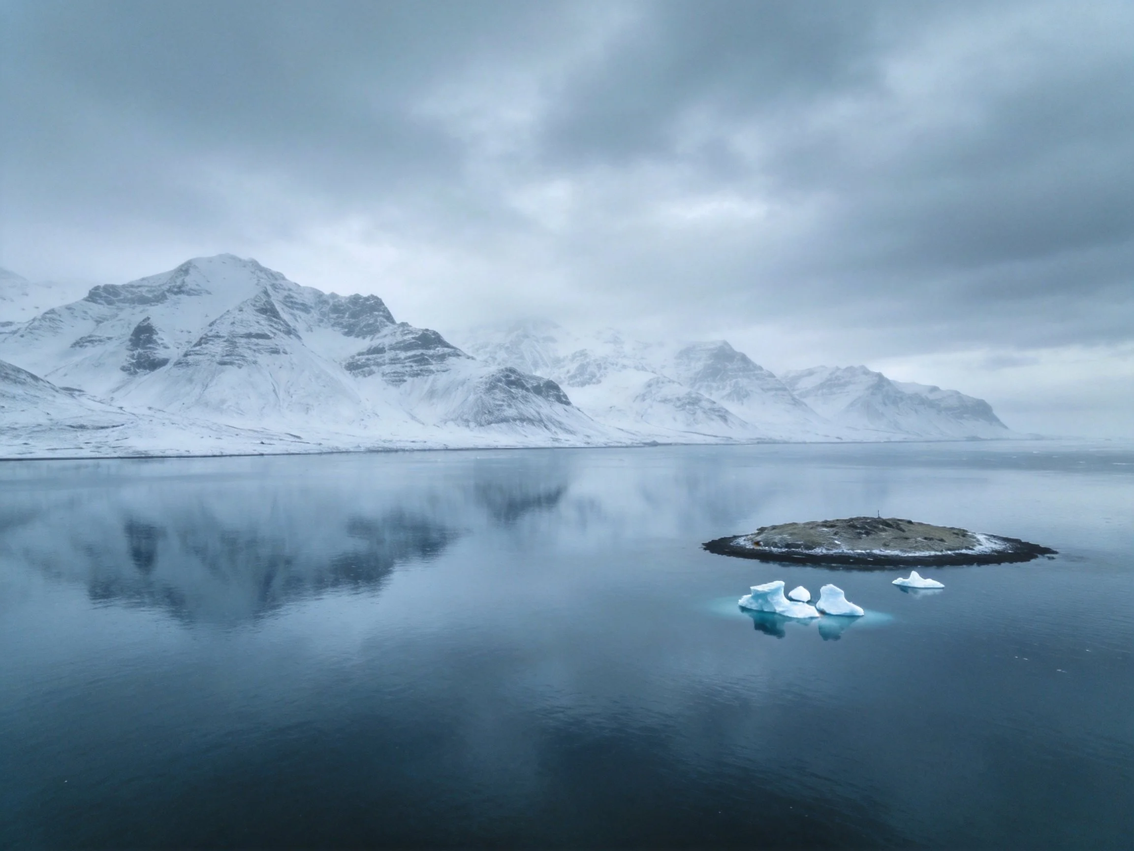 Snow - capped mountain peaks reflect in calm icy waters near rocky islet
