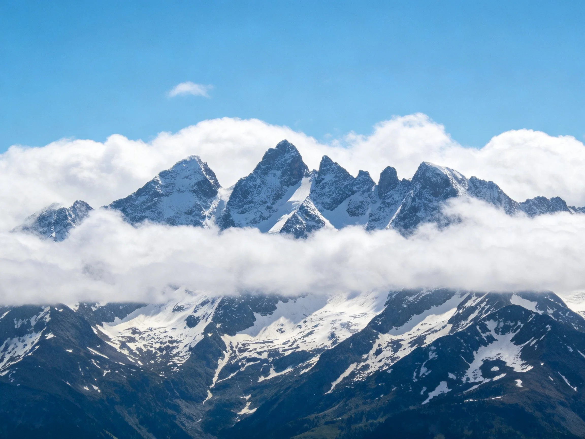 Snow-capped mountain peaks rise through low-hanging clouds against a clear blue sky.