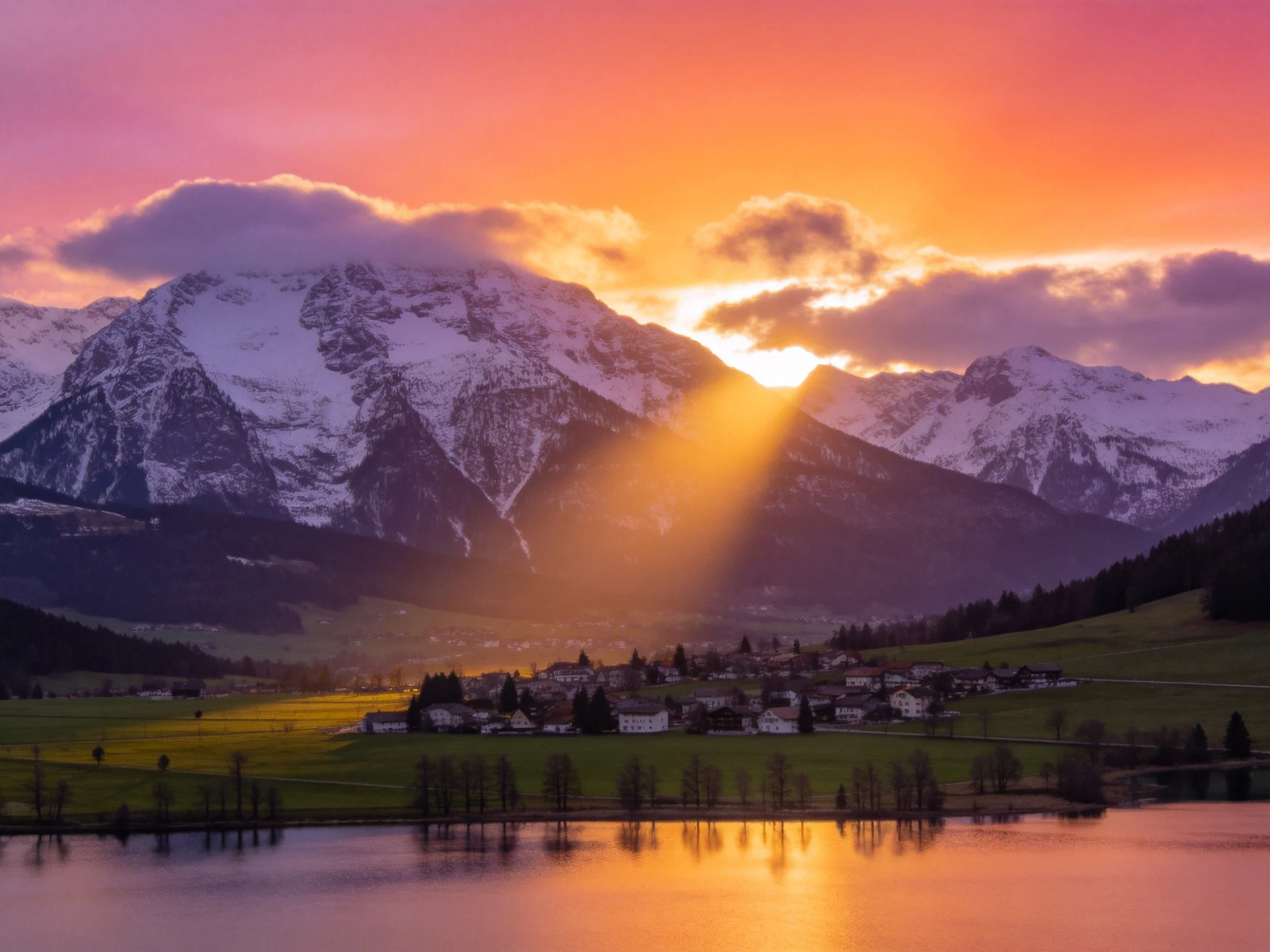 Snow - capped mountains at sunset with village and reflective lake