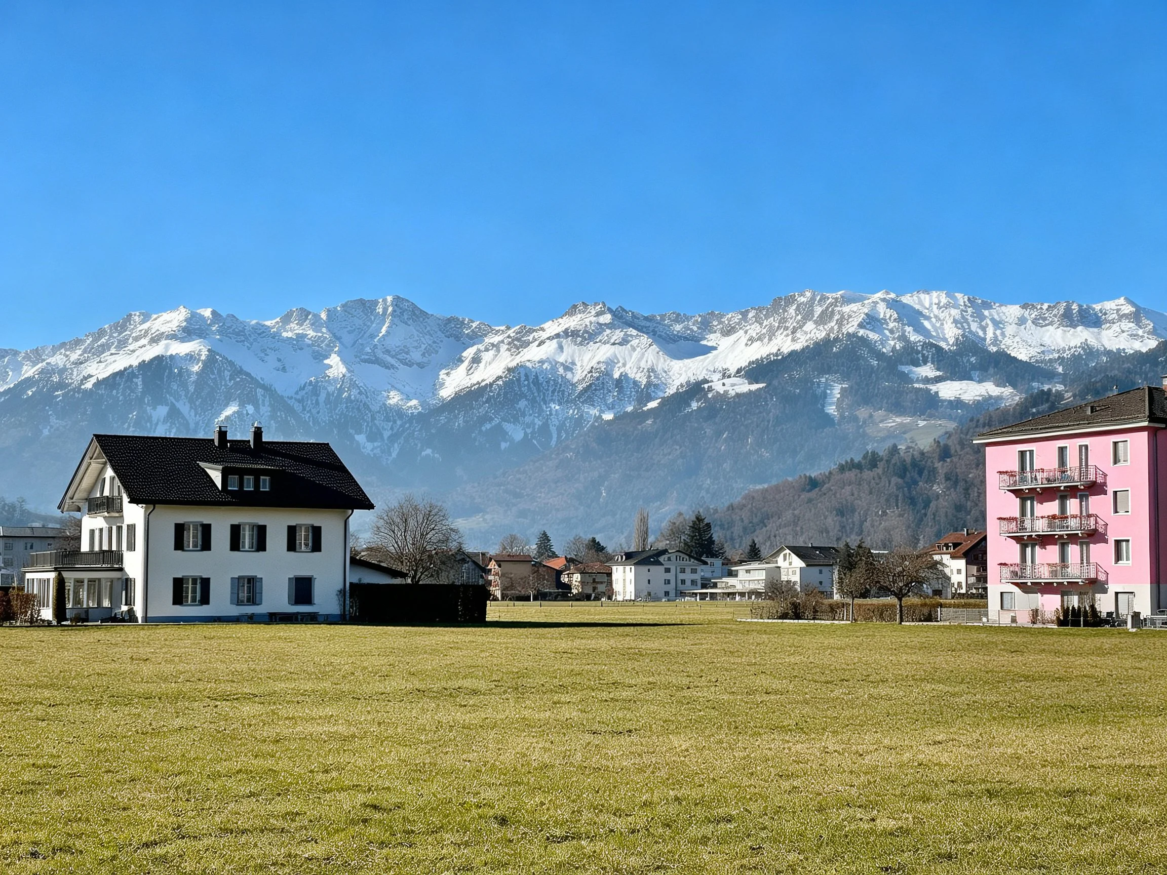 Snow-capped mountains behind white and pink residential buildings over expansive green field
