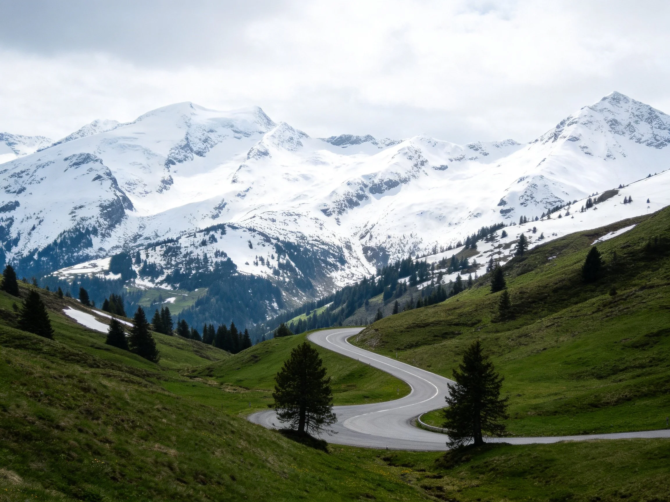 Snow-capped mountains overlook a winding road through lush green hills