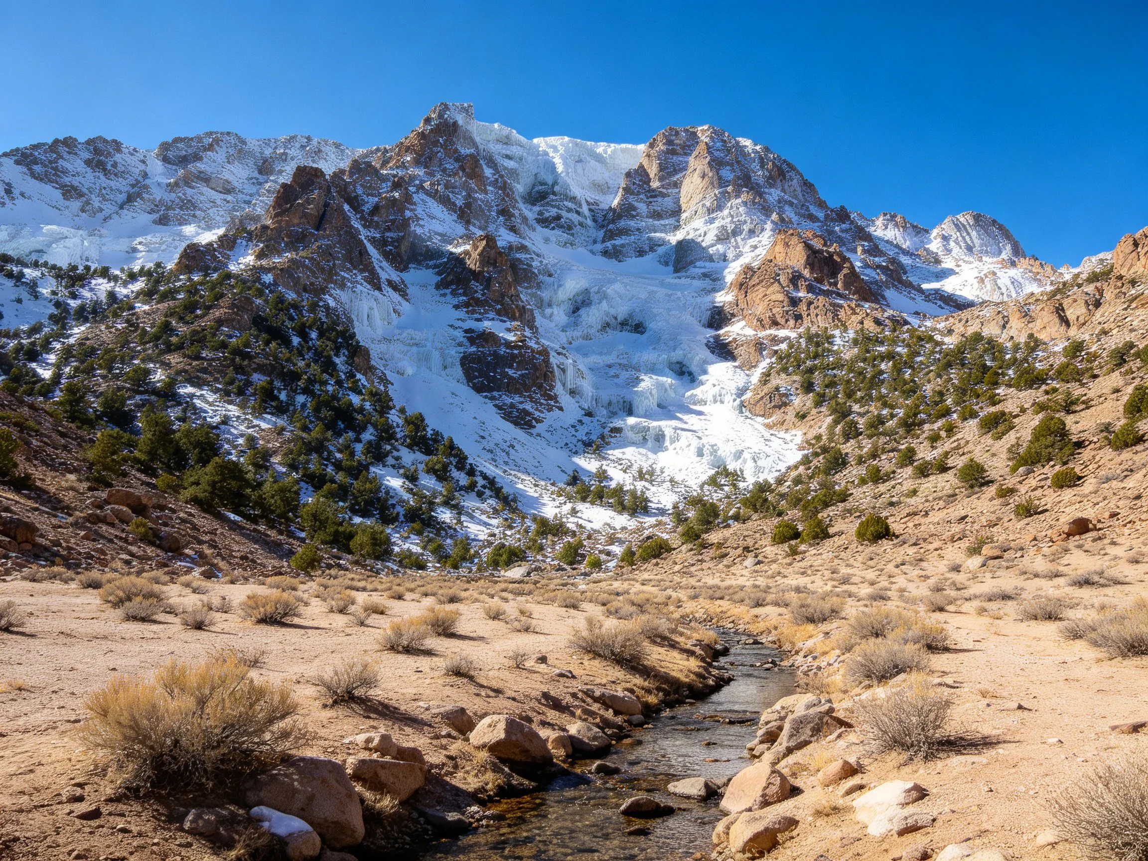 Snow - capped mountains tower over a rugged valley with a narrow stream