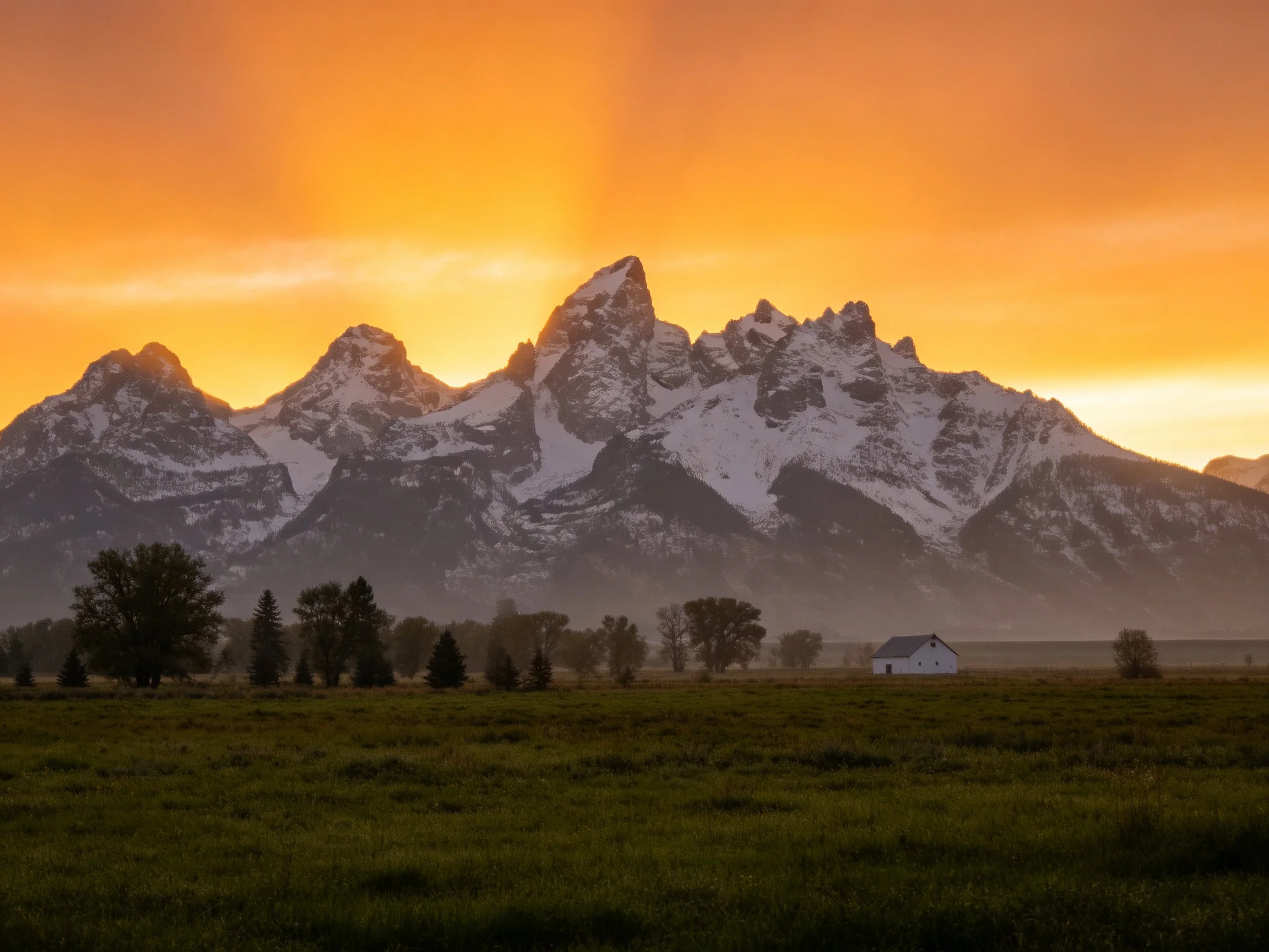 Snow-capped peaks glow under golden sunset sky with rural landscape