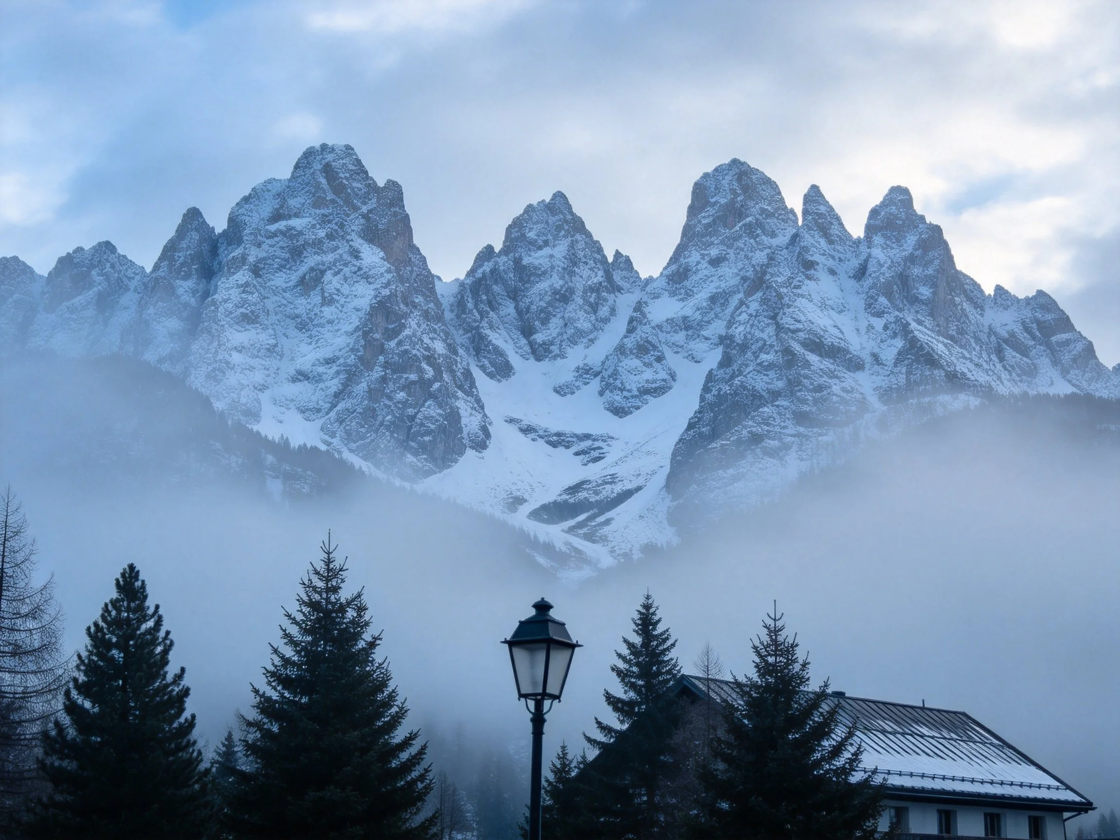 Snow-covered jagged peaks behind misty forest and house
