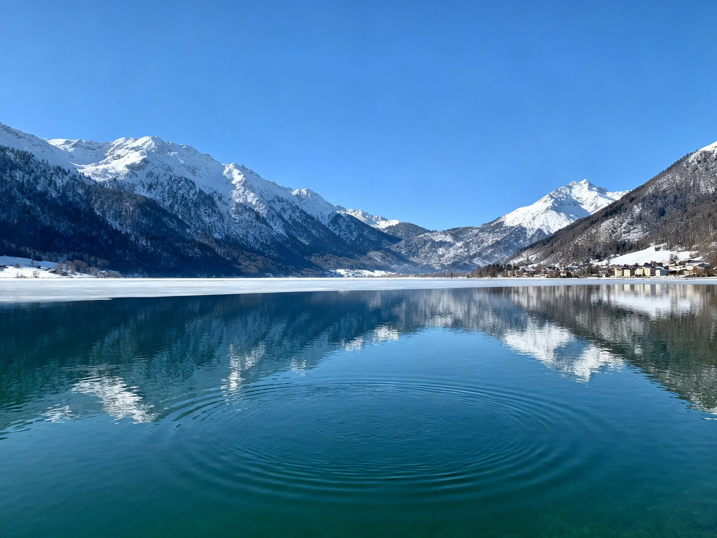 Snow-covered mountain peaks reflect in a frozen lake beneath bright blue sky