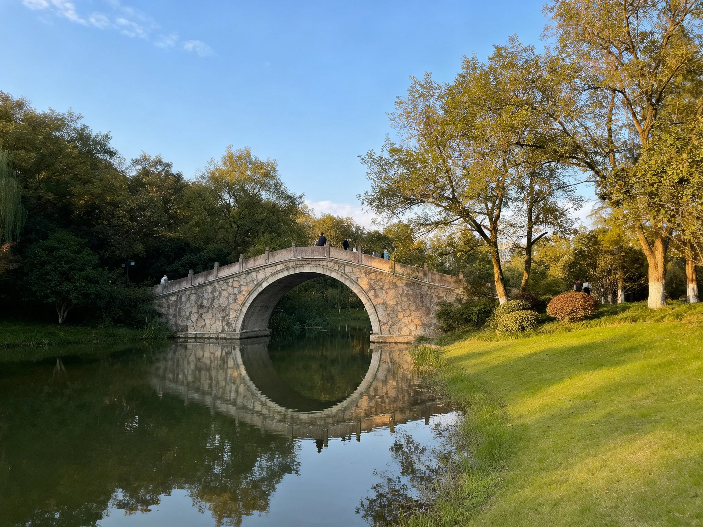 Stone arch bridge spans calm pond with green surroundings
