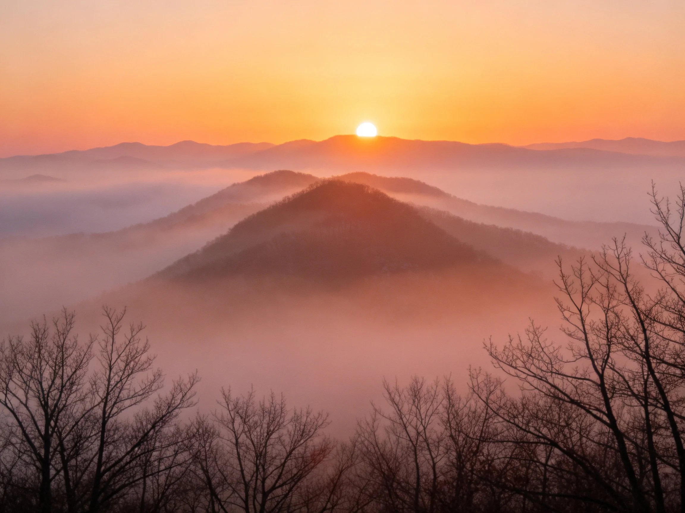 Sunrise over layered mountains with pink-orange haze and silhouetted trees