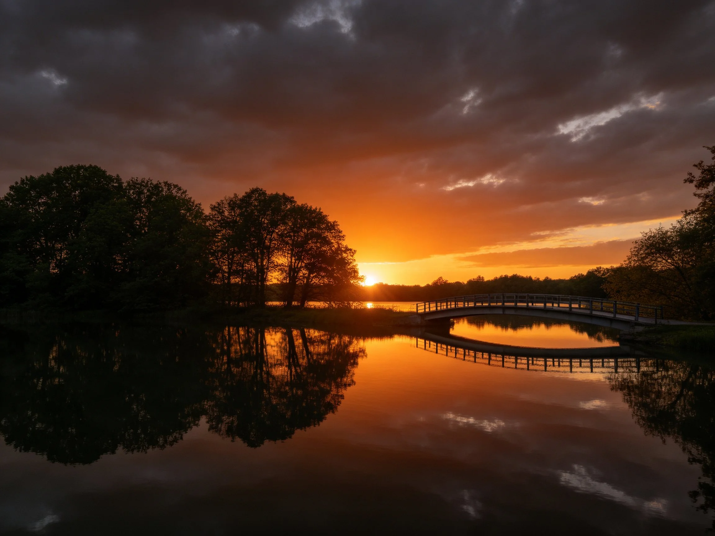 Sunset over calm lake with tree silhouettes and arched bridge