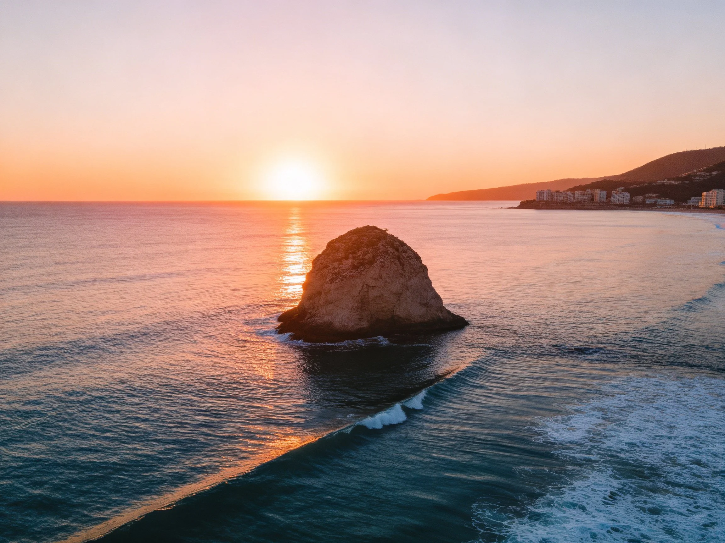 Sunset over calm ocean with central rock formation near coastal buildings