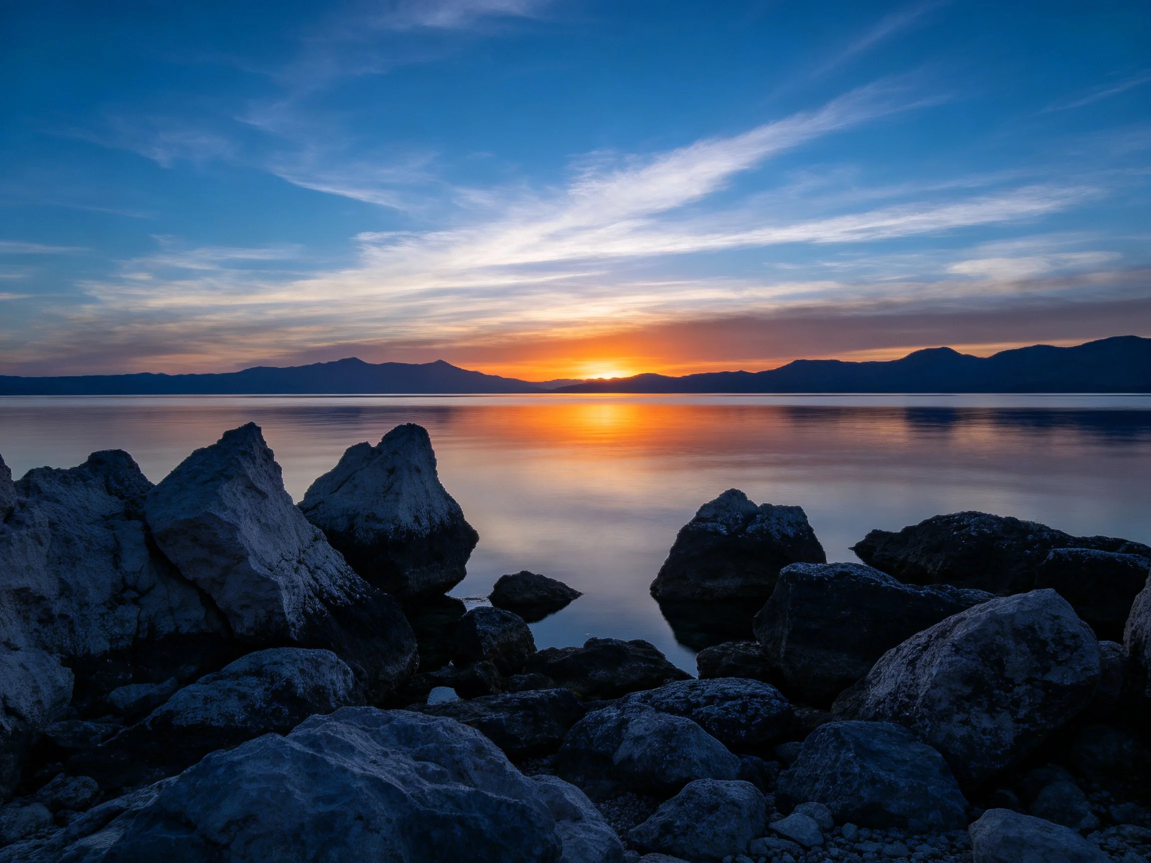 Sunset over calm waters with rocky shoreline and mountain backdrop