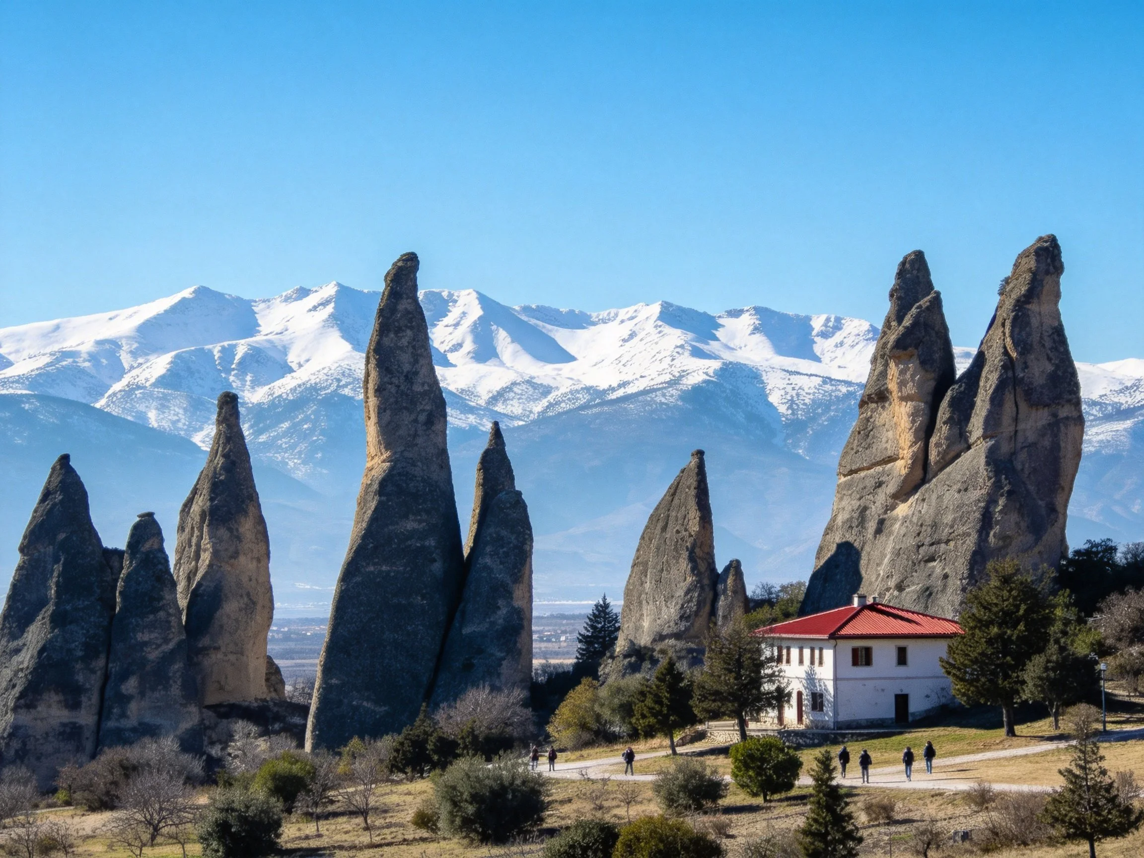 Tall gray rock spires beside white house with red roof, snowy mountain backdrop