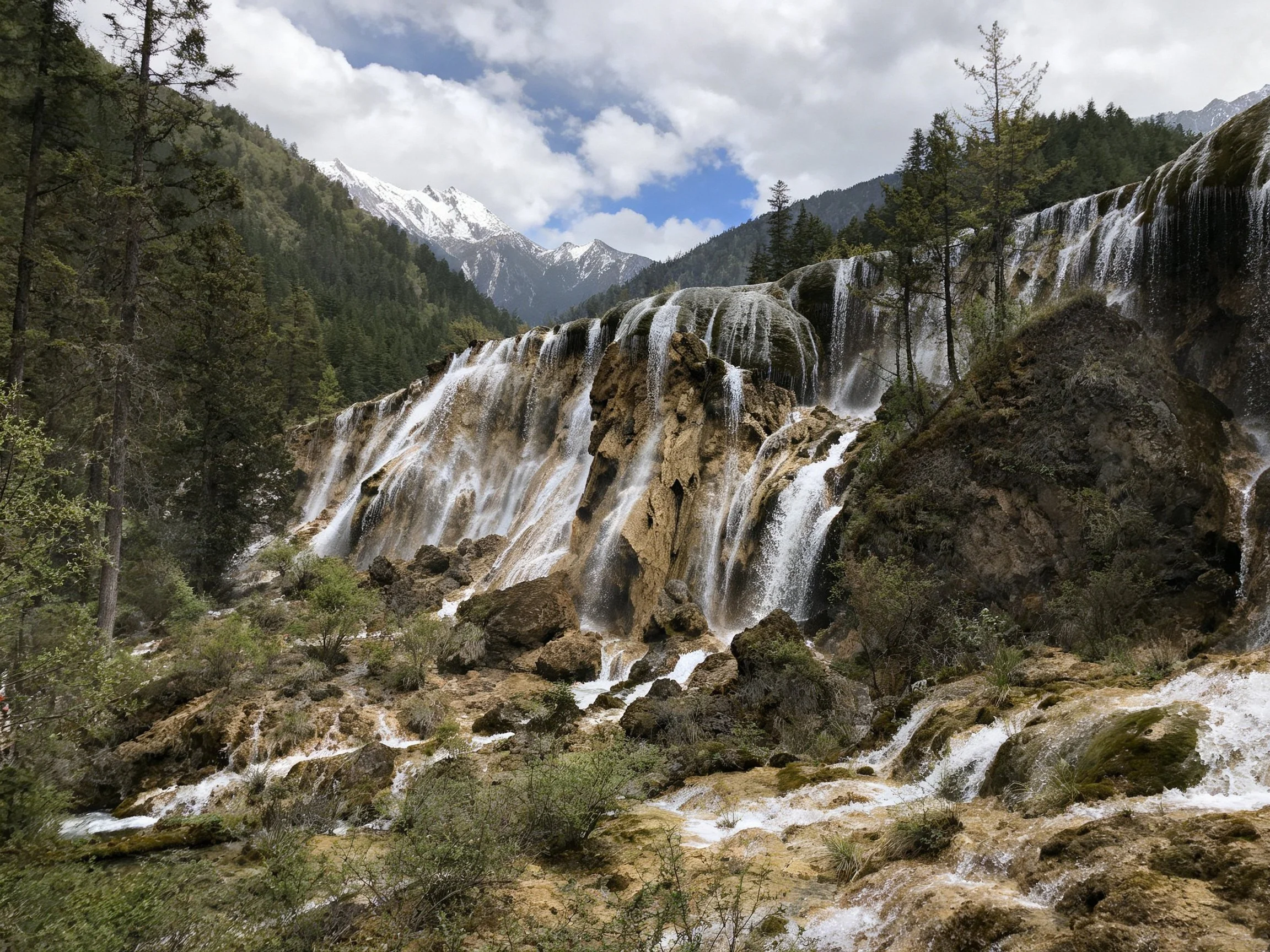 Tiered waterfalls cascade over rocky cliffs amidst dense forests and snow - capped mountains