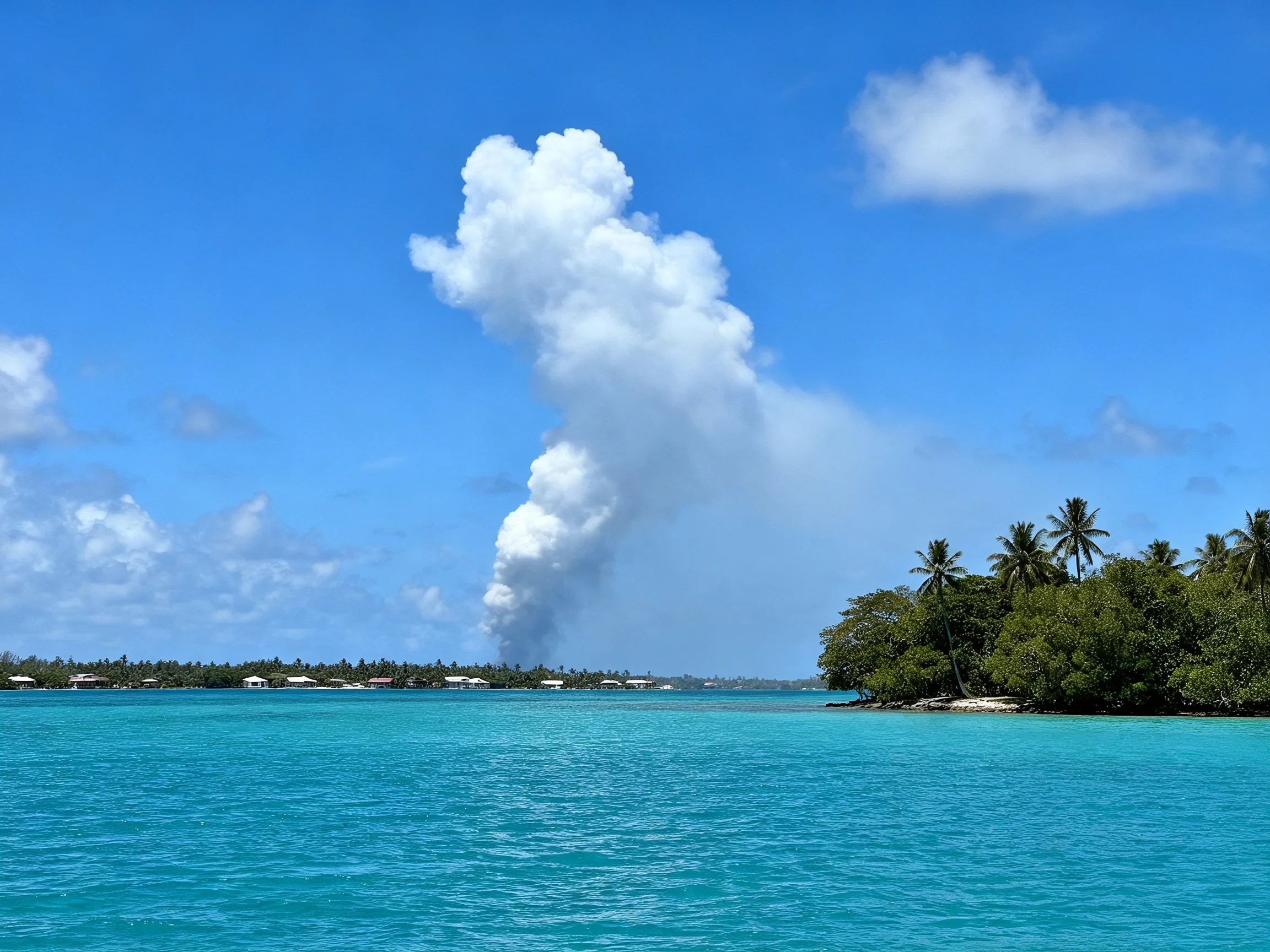 Turquoise water, palm trees, and a large white smoke plume under bright blue sky.