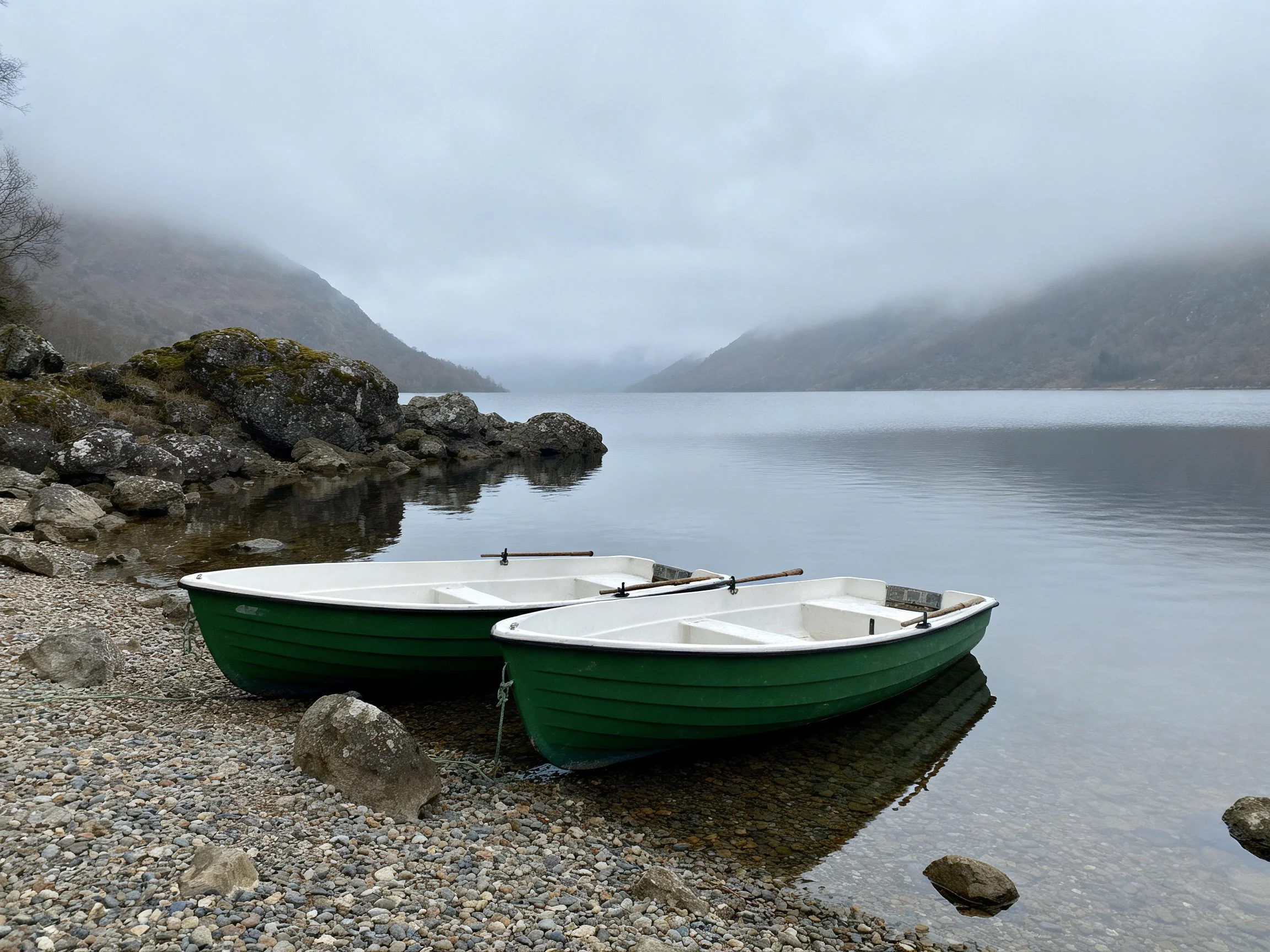 Two green rowboats rest on a pebbly lakeshore with misty mountains
