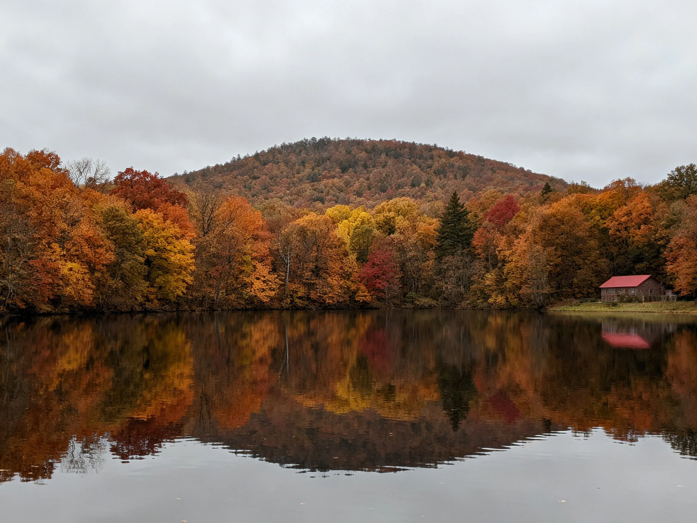 Vibrant autumn trees reflected in calm lake with red-roofed cabin and forested hill
