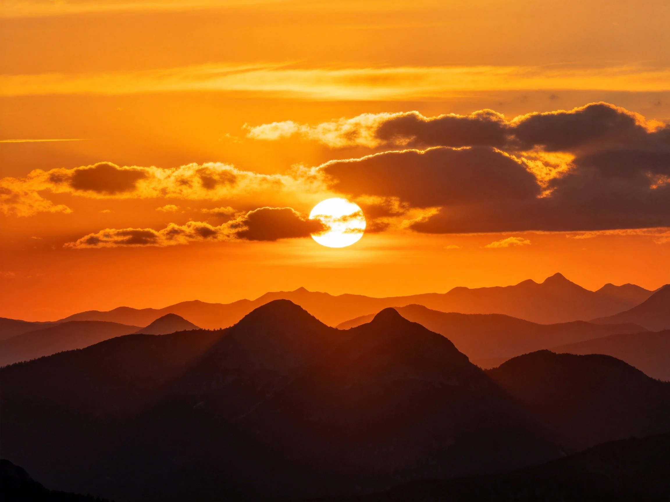 Vibrant orange sunset illuminates layered mountain silhouettes with scattered clouds