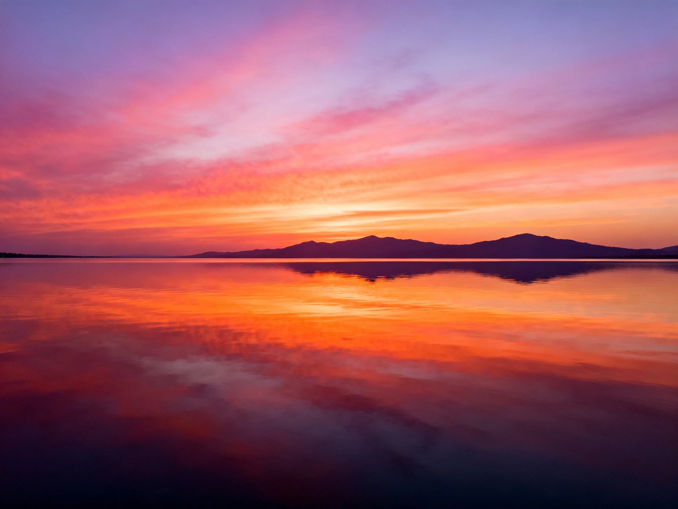 Vibrant pink-orange sunset reflects on calm water near dark mountains.
