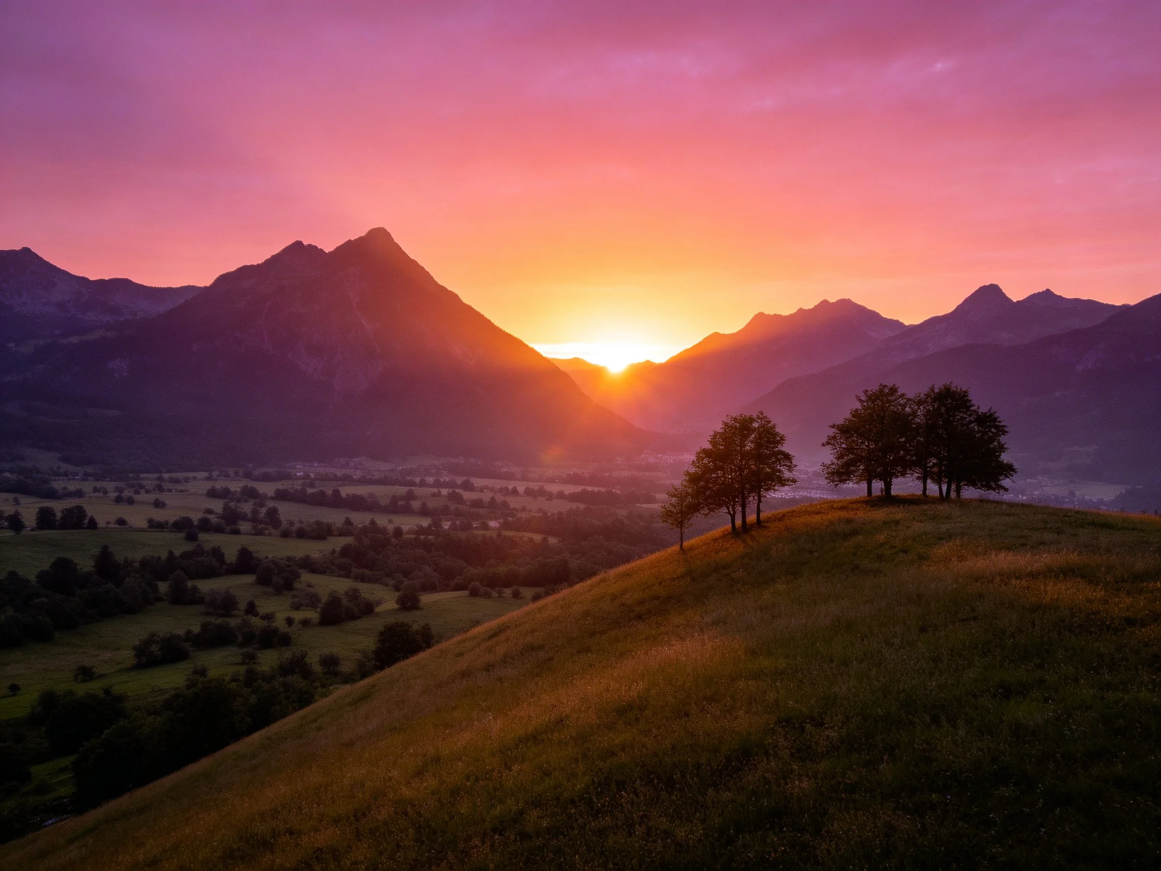 Vibrant sunset illuminates mountain peaks with silhouetted trees on a grassy hill