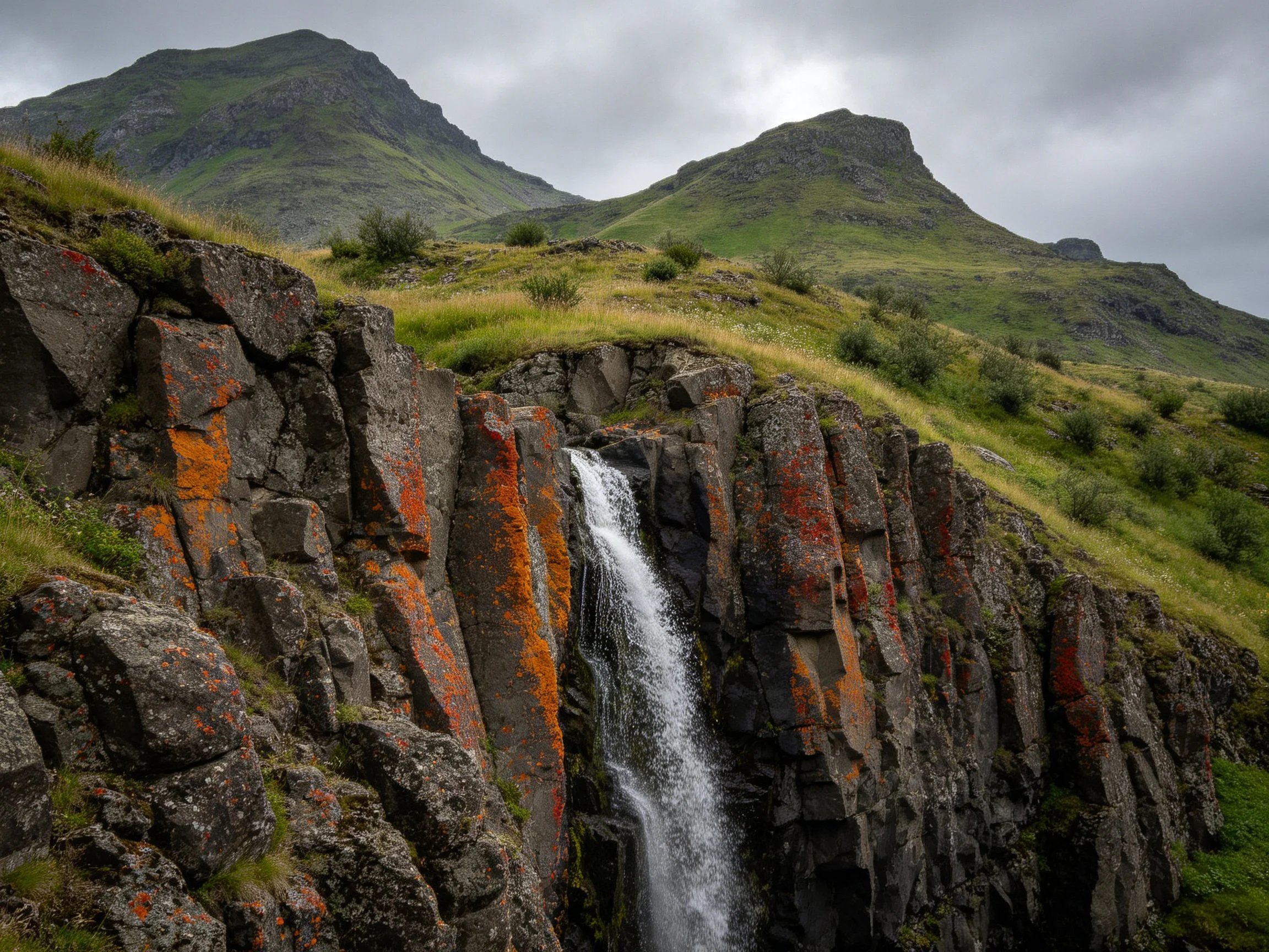 Waterfall over rocky cliff with orange lichen and green hills