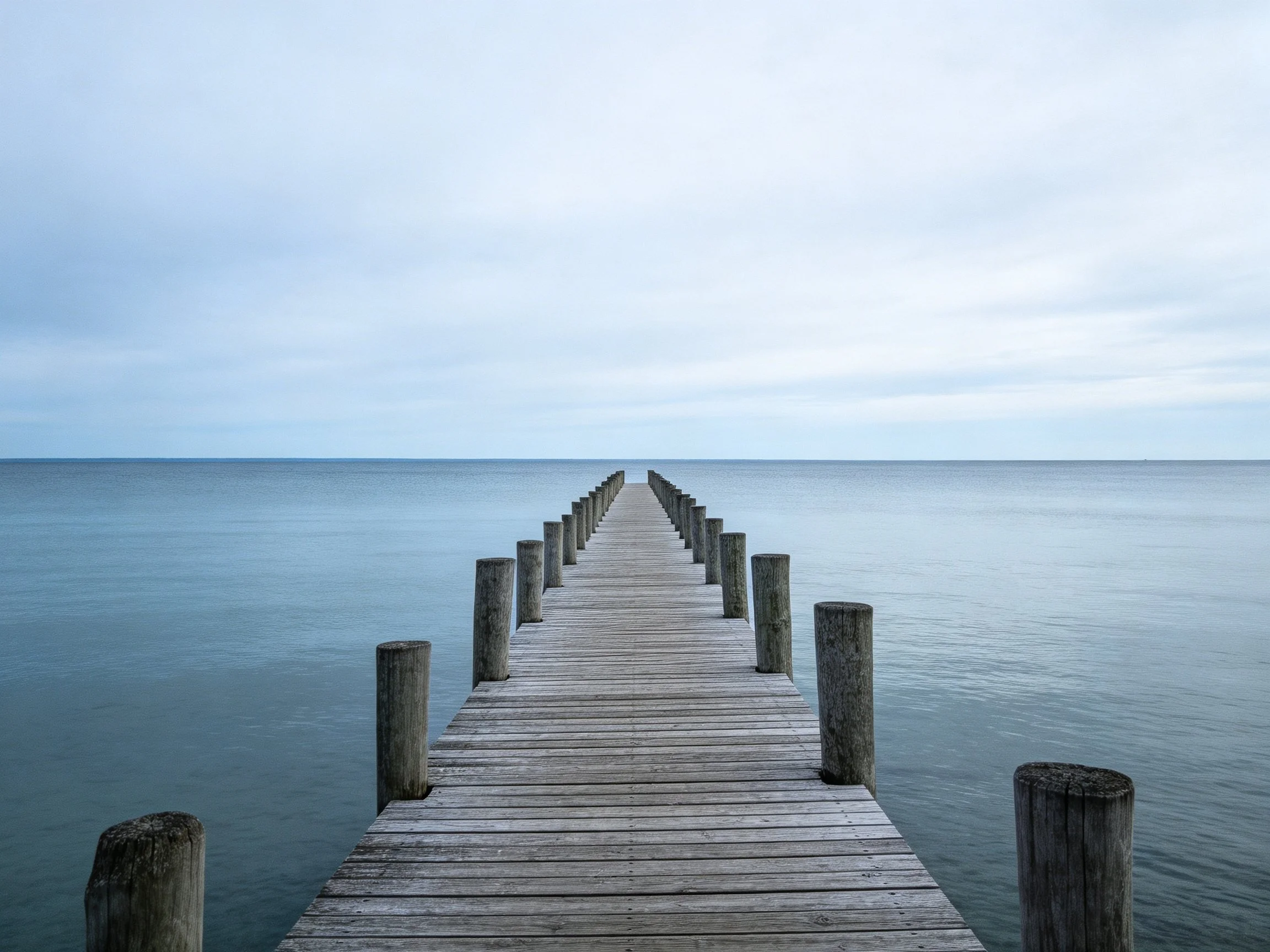 Weathered wooden pier stretches into calm blue water under overcast sky