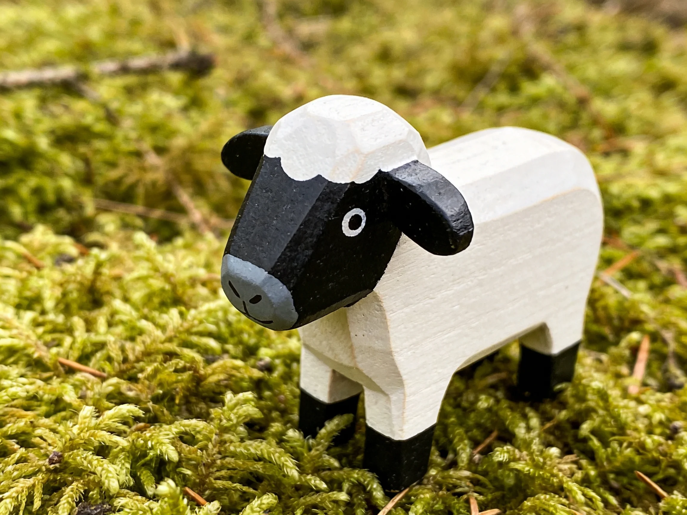 Wooden sheep figurine with black face and white wool on mossy ground
